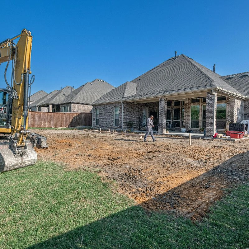 A yellow excavator grading a dirt backyard area behind a large brick residential home in Anaheim Hills.