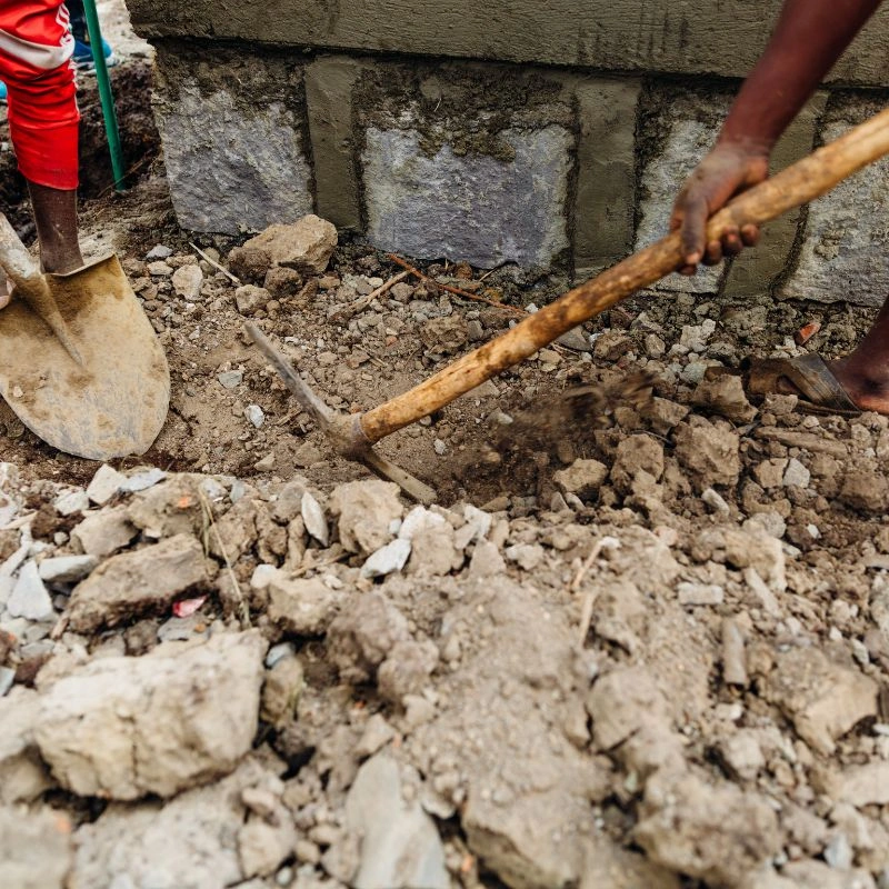 A close shot of a worker utilizing a pickaxe to break up compacted rocky soil beneath a stone footing in Tustin.