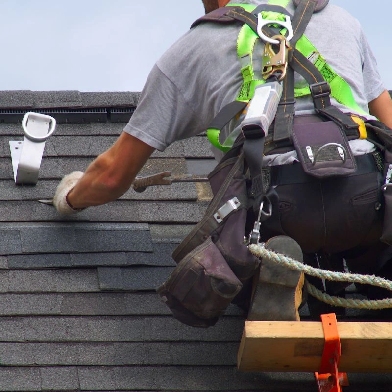 A roofer in a high-elevation harness using a pry bar near a newly mounted metal safety anchor in Fullerton.