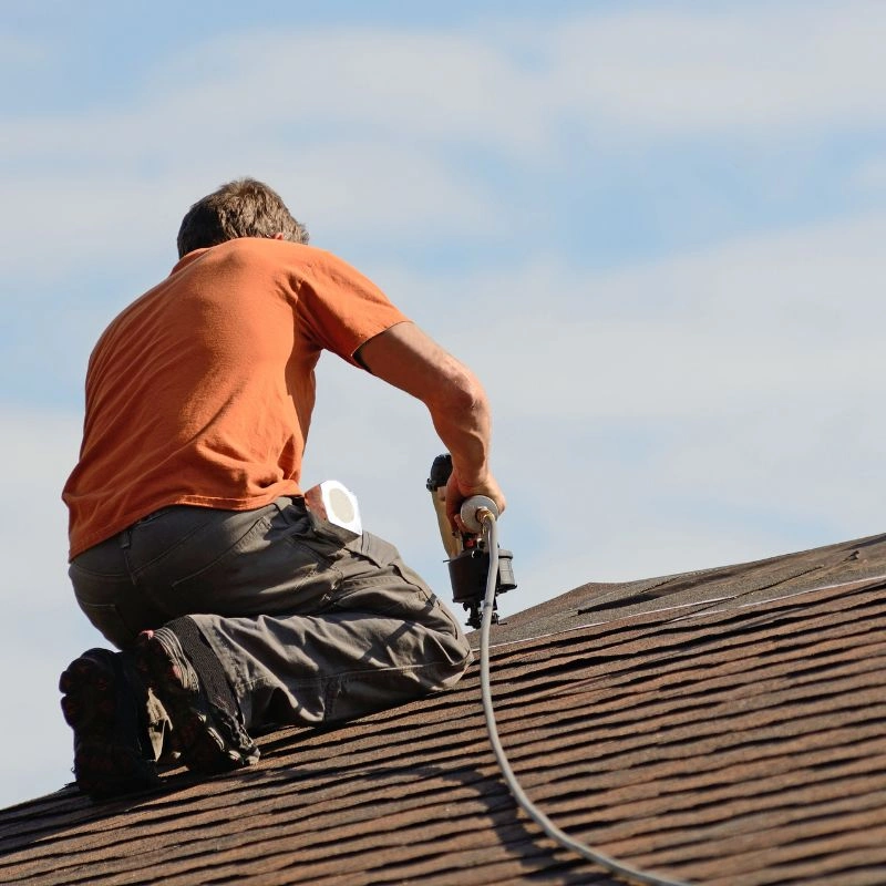 A roofer kneeling on a residential slope while using a pneumatic coil nail gun on architectural shingles in Tustin.